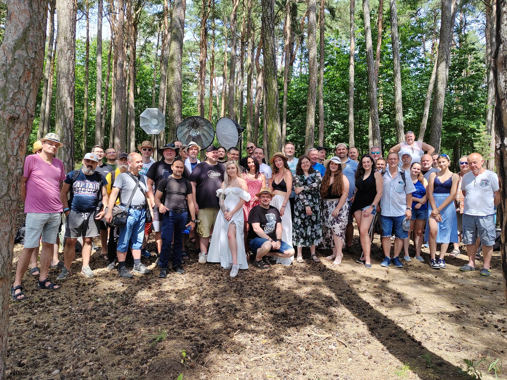 Velká skupina lidí pózuje pro fotografii v zalesněné oblasti s osvětlovacím zařízením viditelným v pozadí za slunečného dne.